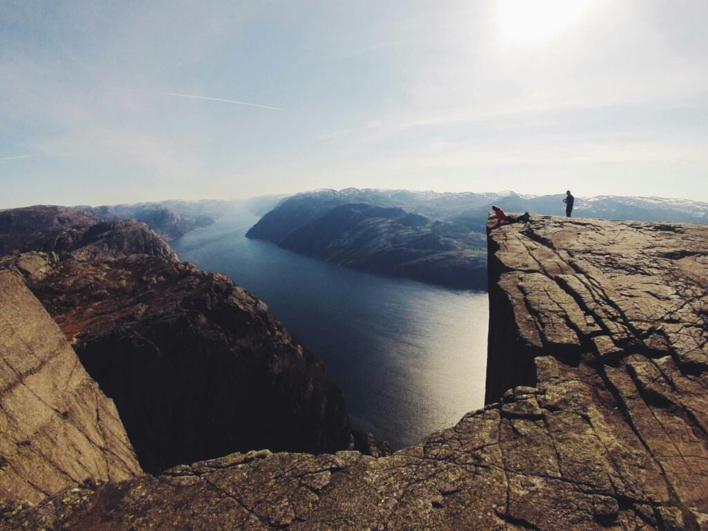 Silhouette of adventurers on a dramatic cliff overlooking a serene fjord under clear skies.