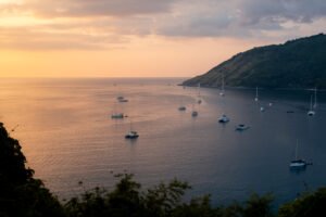 Tranquil view of sailboats and yachts in Phuket, Thailand with a warm sunset horizon.