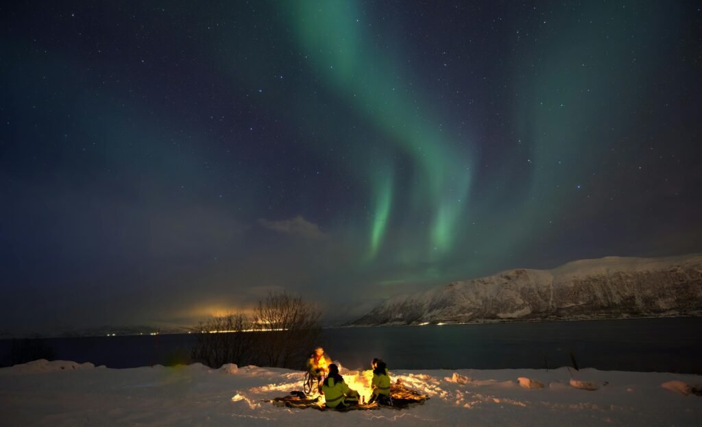 Two people enjoy the Aurora Borealis by a campfire near Tromsø, Norway's snowy landscape.
