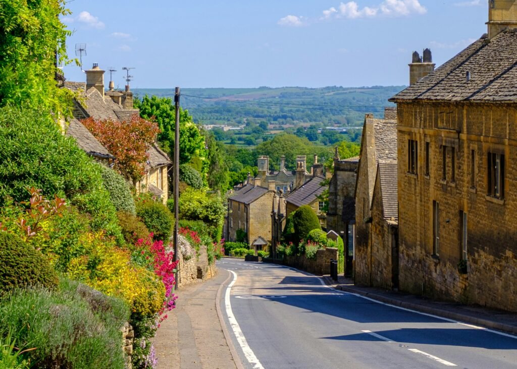 Picturesque view of a winding street with stone houses and lush greenery in a Cotswold village, England.
