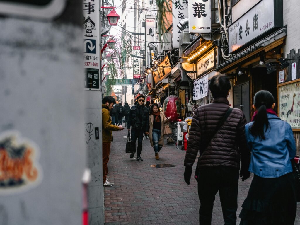 A lively street scene featuring people walking by shops with Japanese signage in a busy alleyway.